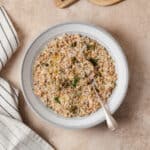 Lentil dip shown in a grey bowl with white strip along the edge. Spoon resting in the dip on the right side of the bowl. On a beige background with a black and white striped napkin on the left.