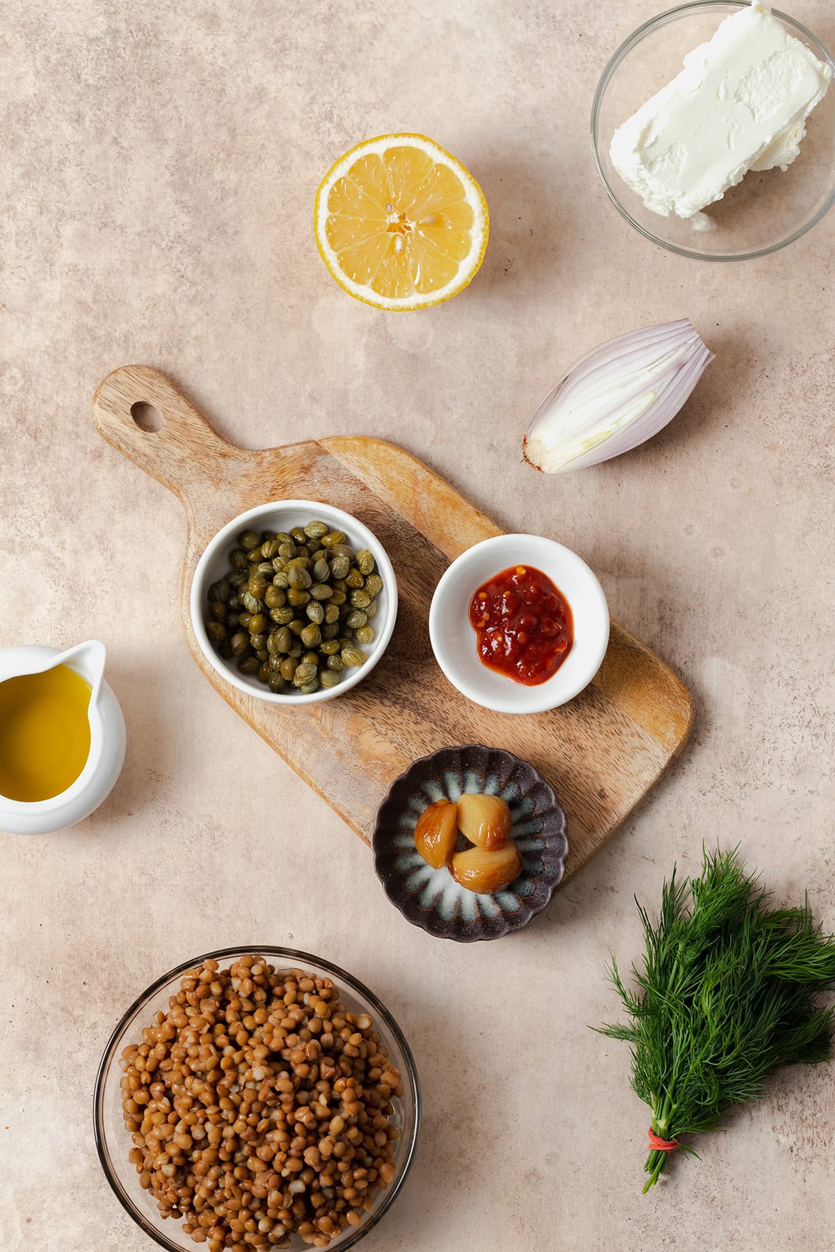 Ingredients laid out on a beige background. Cooked brown lentils, capers, olive oil, half a lemon, a block of soft goat cheese, half a shallot, a bunch of fresh dill, roasted garlic, and garlic chili sauce.