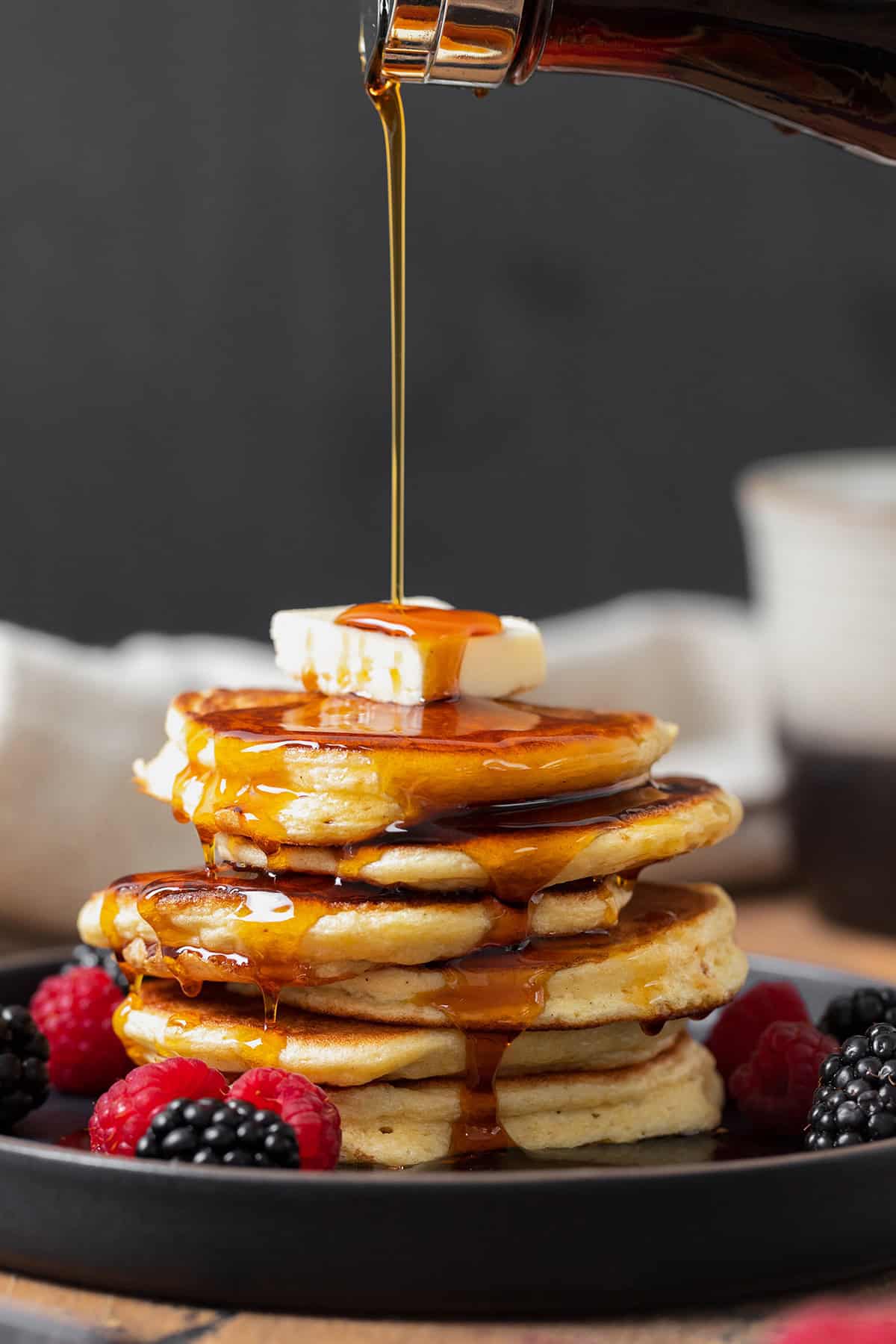 Pancakes shown stacked on a black plate with butter on top and maple syrup being poured on.