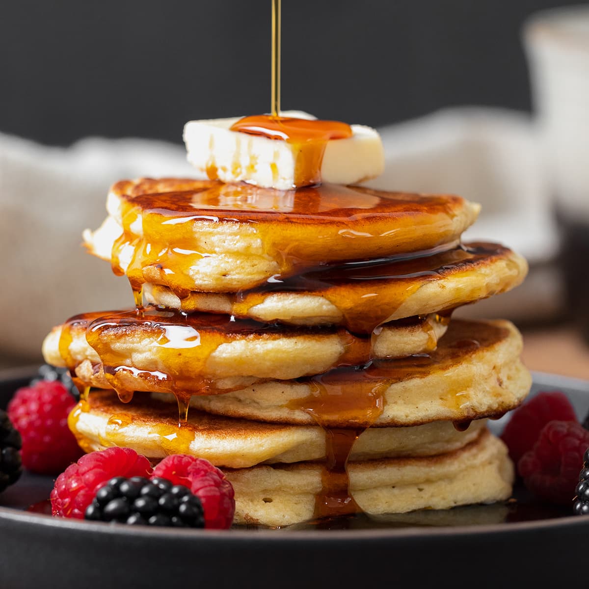 Pancakes shown stacked on a black plate with butter on top and maple syrup being poured on.