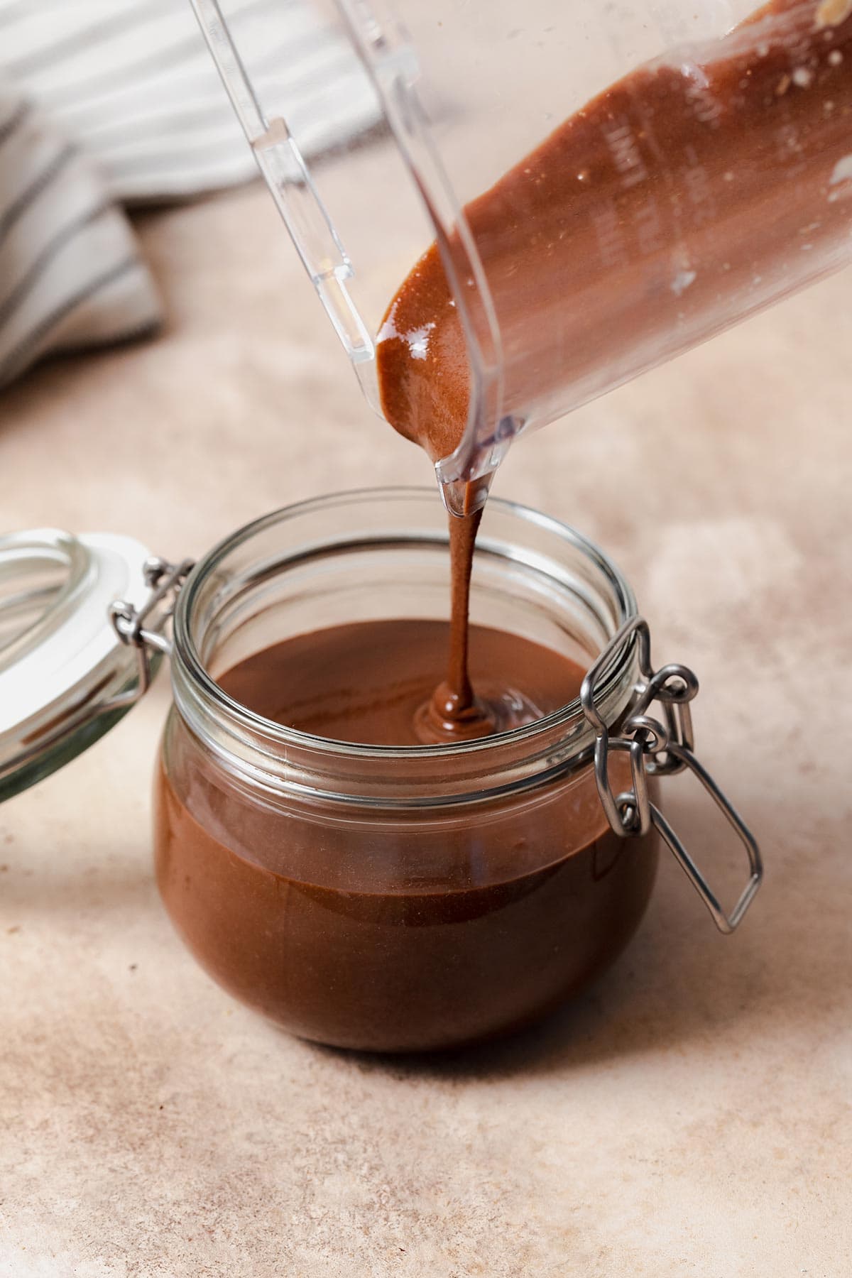 A pouring shot of healthy nutella being poured from the blender into a glass jar. On a beige background.