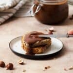 Healthy Vegan Nutella spread on toasted bread. On a white plate and beige background. A butterknife spreading more nutella on the bread from the right.