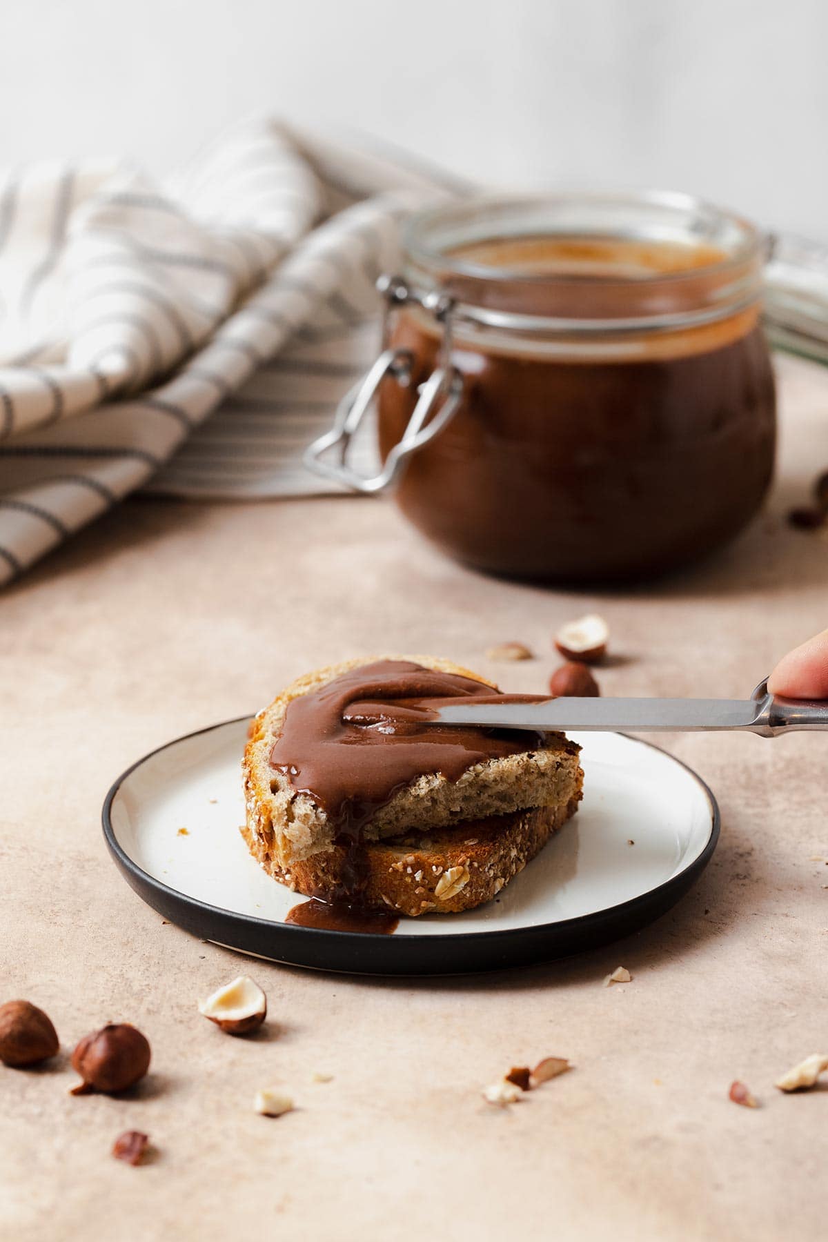 Healthy Vegan Nutella spread on toasted bread. On a white plate and beige background. A butterknife spreading more nutella on the bread from the right.