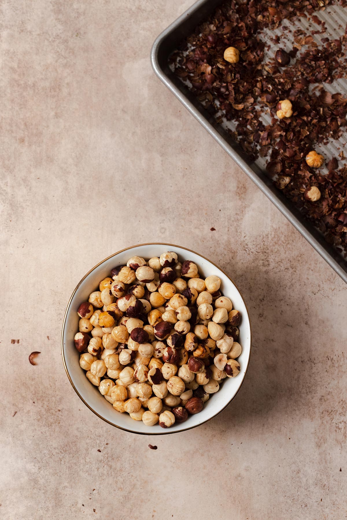 Roasted hazelnuts in a white bowl on a beige background with the baking sheet in the top right corner.