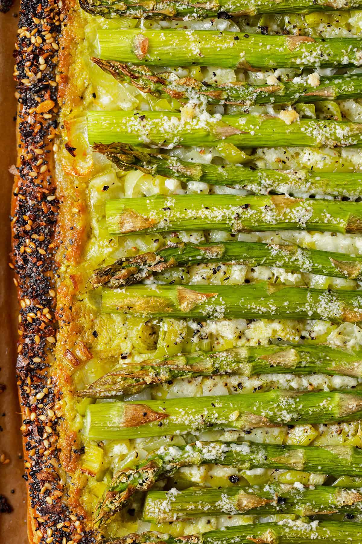 A close up of the filling and the left edge of the tart. Asparagus, goat cheese pesto spread, pecorino cheese with golden bits from baking. Crust sprinkled with everything bagel seasoning.