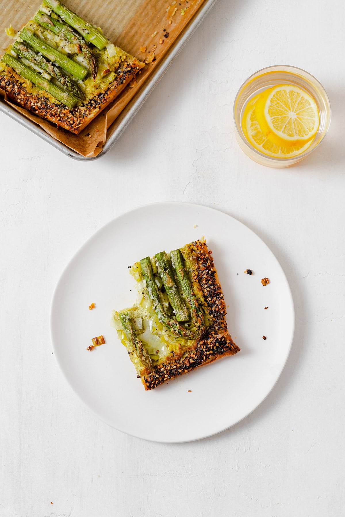 Asparagus tart square with a bite taken out of it on a while plate in the middle, a glass of water with a slice of lemon on the top right, and the pan with the rest of the tart in the top left corner.