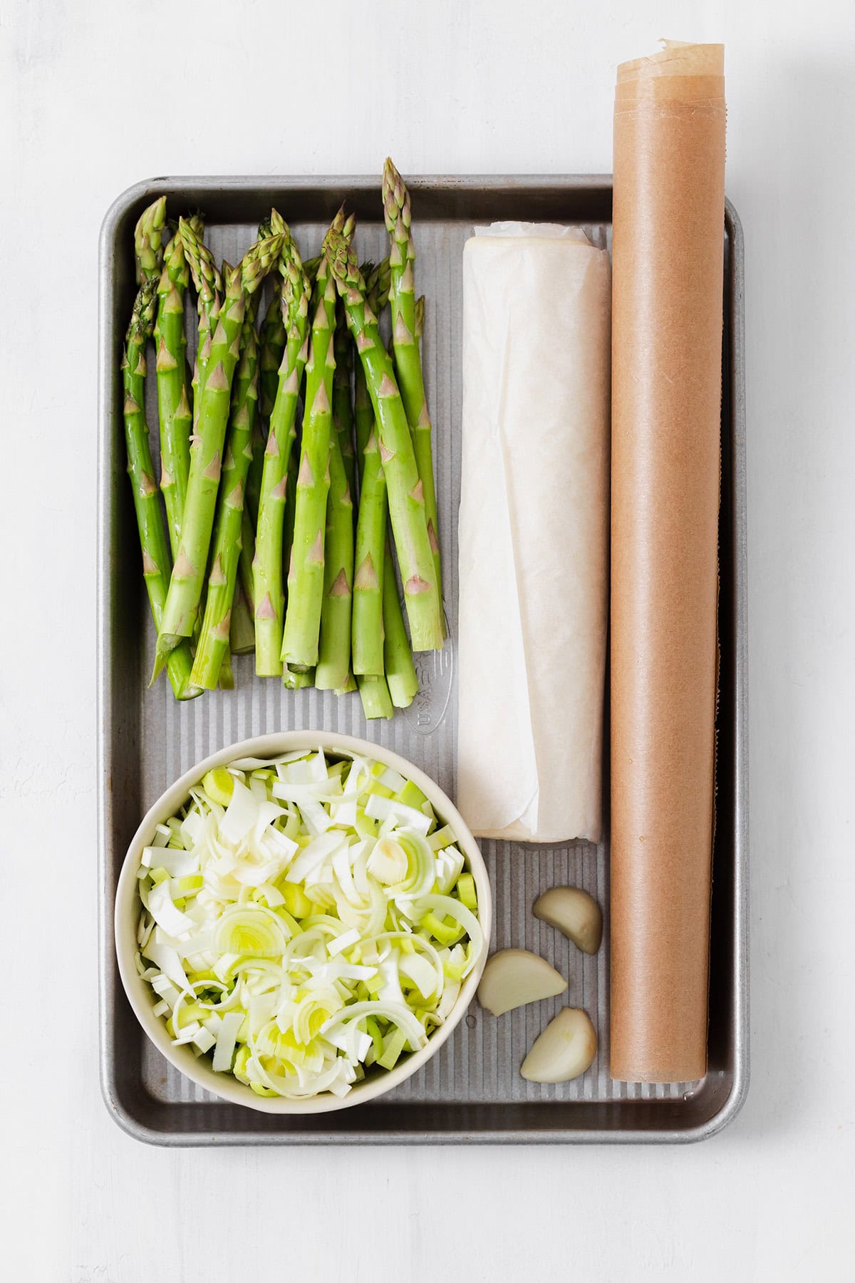 Ingredients for asparagus tart laid out on a baking sheet - asparagus, chopped leek, garlic cloves, puff pastry, and parchment paper