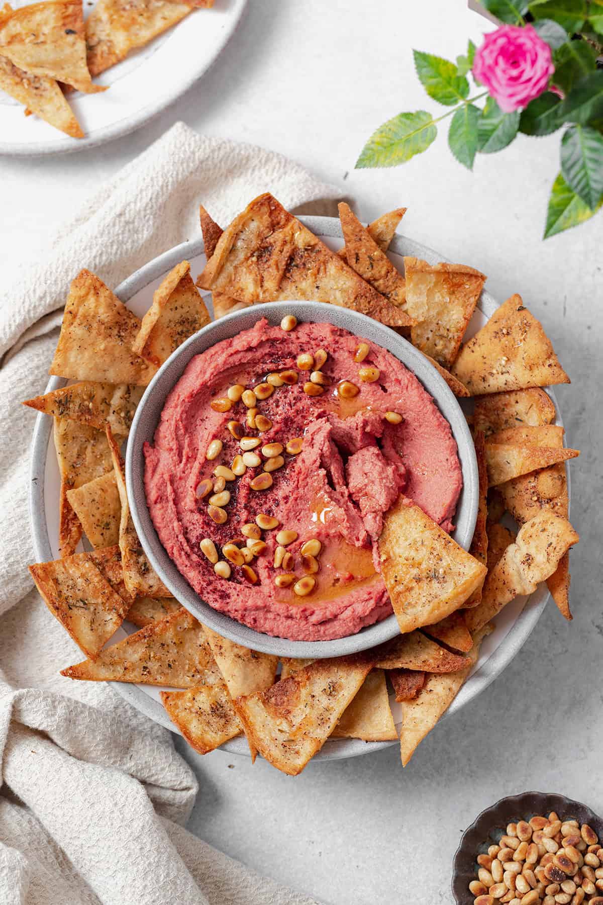 Beet hummus shown in a grey bowl on a grey plate surrounded by homemade pita chips. Grey background with a beige tea towel on the left of the bowl. One pita chip dipped in the hummus on the right side.