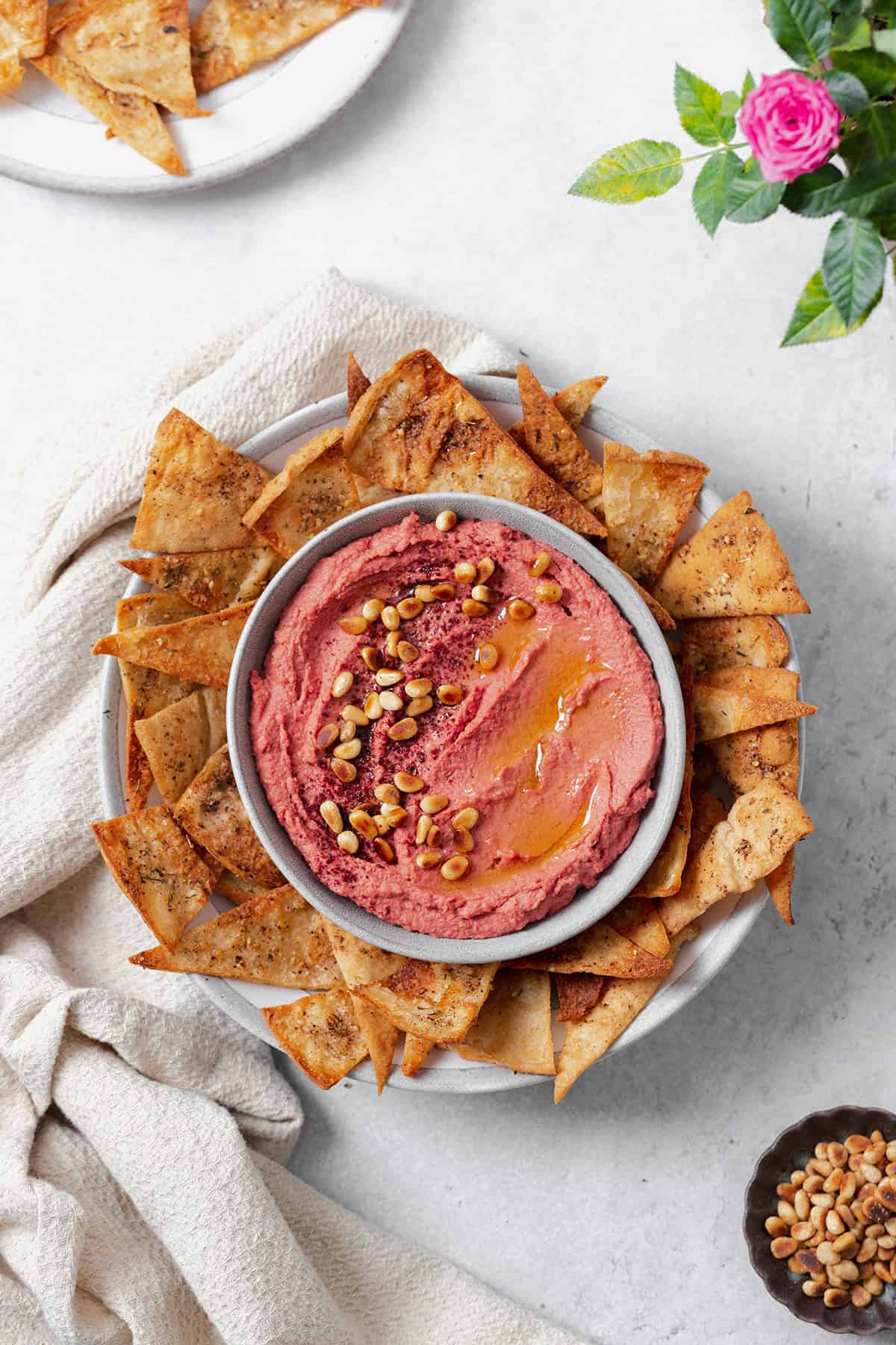 Beet hummus shown in a grey bowl on a grey plate surrounded by homemade pita chips. Grey background with a beige tea towel on the left of the bowl. One pita chip dipped in the hummus on the right side. More chips on a plate in the top left corner, a rose in the top right corner, and pine nuts in the bottom right corner.