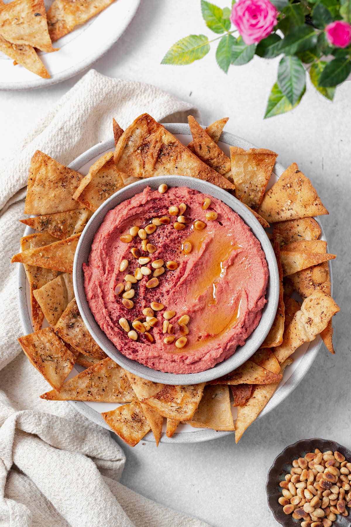Beet hummus shown in a grey bowl on a grey plate surrounded by homemade pita chips. Grey background with a beige tea towel on the left of the bowl. One pita chip dipped in the hummus on the right side. More chips on a plate in the top left corner, a rose in the top right corner, and pine nuts in the bottom right corner.