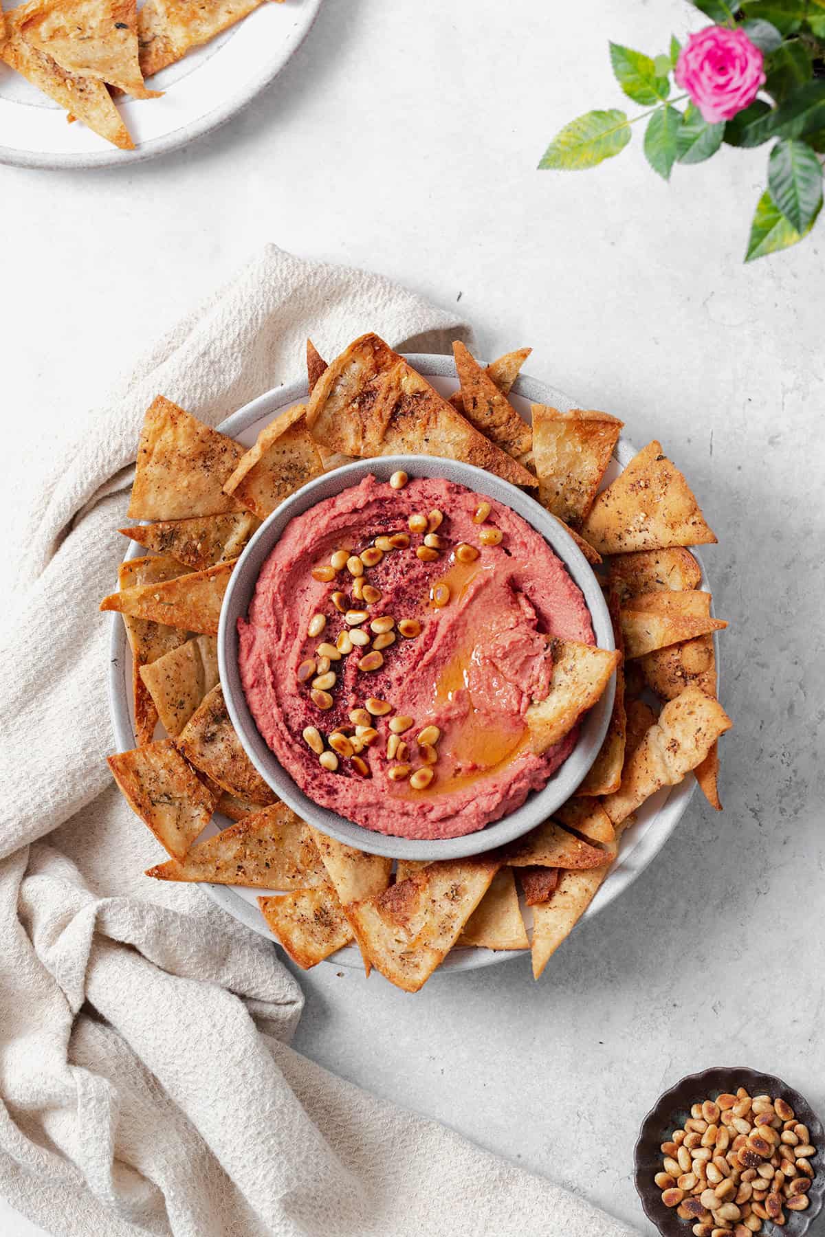 Beet hummus shown in a grey bowl on a grey plate surrounded by homemade pita chips. Grey background with a beige tea towel on the left of the bowl. One pita chip dipped in the hummus on the right side. More chips on a plate in the top left corner, a rose in the top right corner, and pine nuts in the bottom right corner.