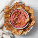 Beet hummus shown in a grey bowl on a grey plate surrounded by homemade pita chips. Grey background with a beige tea towel on the left of the bowl.
