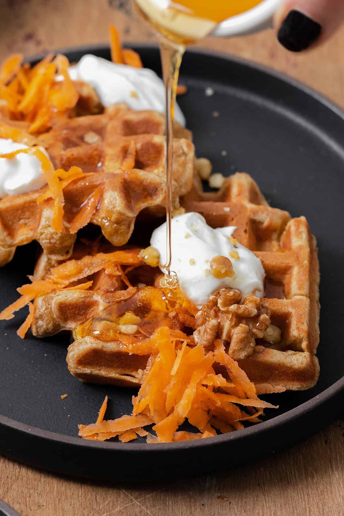 A photo of a hand holding a small bowl of honey and pouring it over two carrot cake waffles on a black plate topped with grated carrots, dollops of greek yogurt, and walnuts.