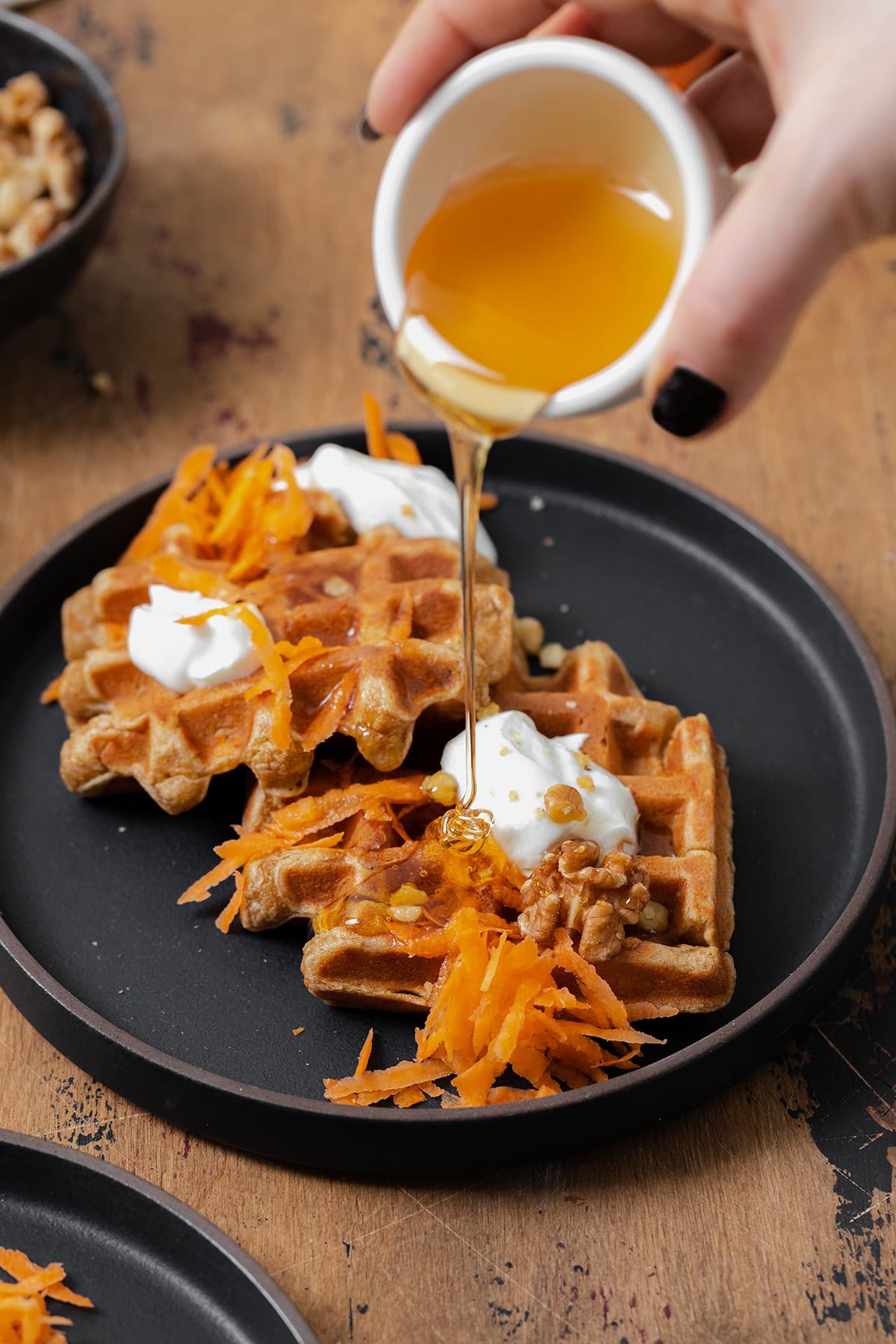 A photo of a hand holding a small bowl of honey and pouring it over two carrot cake waffles on a black plate topped with grated carrots, dollops of greek yogurt, and walnuts.