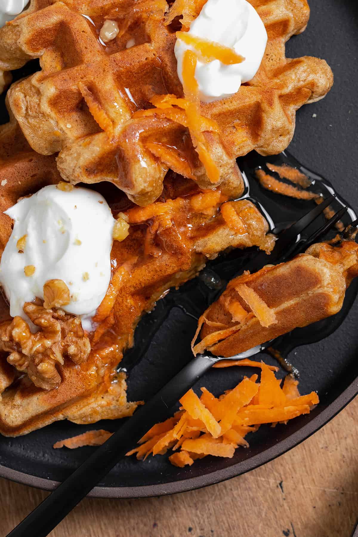 A close up photo of two carrot cake waffles shown on a black plate topped with greek yogurt, honey, grated carrots, and walnuts. A black fork cut a bite-sized piece from one waffle.