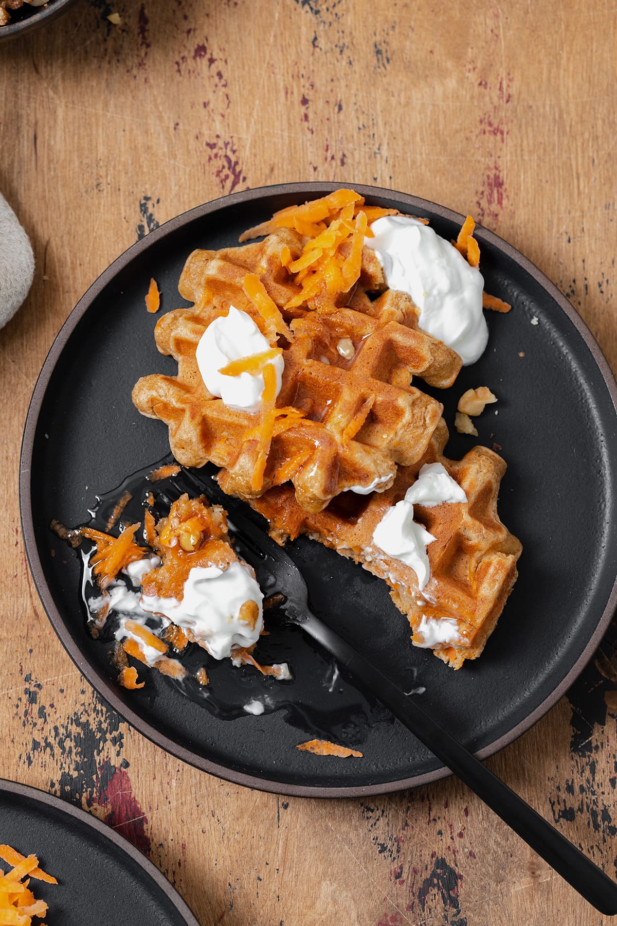 Two carrot cake waffles shown on a black plate topped with greek yogurt, honey, grated carrots, and walnuts. A black fork cut a bite-sized piece from one waffle.