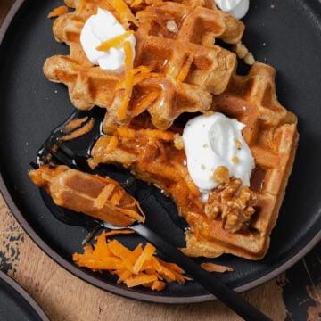 Two carrot cake waffles shown on a black plate topped with greek yogurt, honey, grated carrots, and walnuts. A black fork cut a bite-sized piece from one waffle.