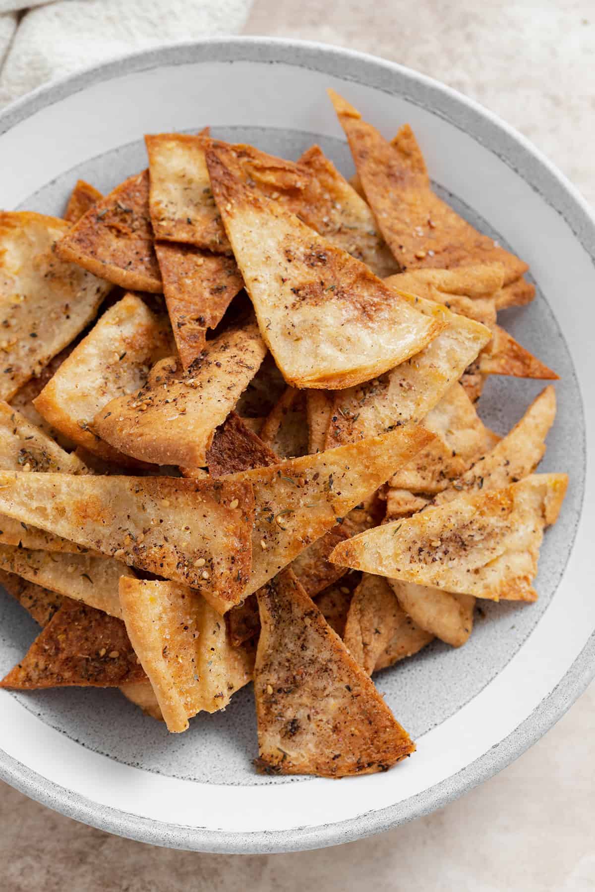 A close up photo of homemade pita chips in a grey bowl on a beige background.