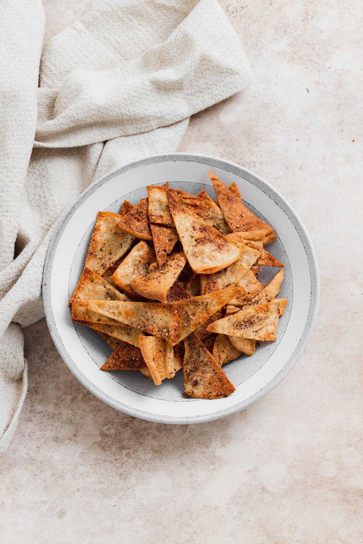 Homemade pita chips in a grey bowl on a beige background with a beige tea towel in the top left corner.