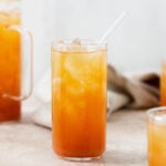 Iced Guava Black Tea shown in a tall glass with ice and a glass straw on beige background. More glasses and a jug full of iced tea partially in the frame on both left and right. Beige tea towel blurry in the background.