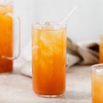 Iced Guava Black Tea shown in a tall glass with ice and a glass straw on beige background. More glasses and a jug full of iced tea partially in the frame on both left and right. Beige tea towel blurry in the background.