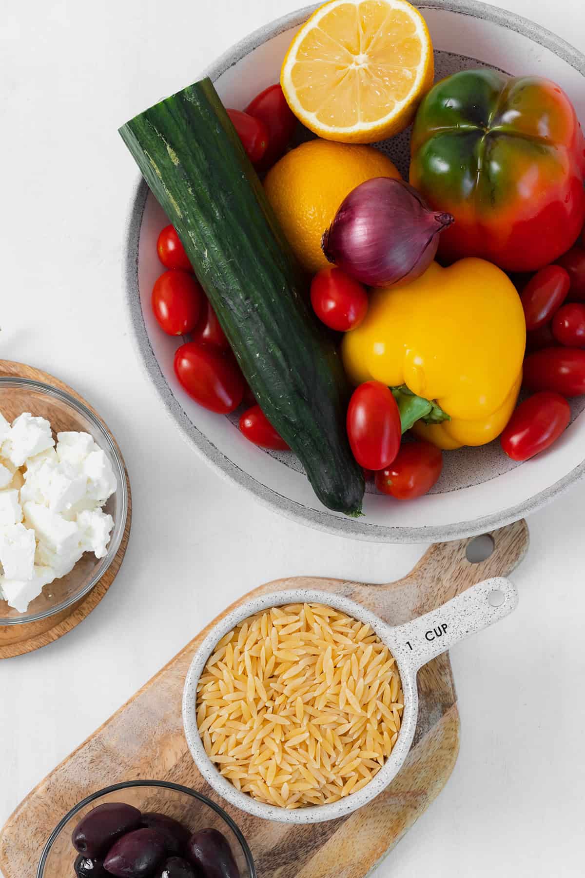 Ingredients for the orzo salad shown laid out on a white background. Cucumber, tomatoes, bell peppers, lemons, feta cheese, orzo, and red onion.