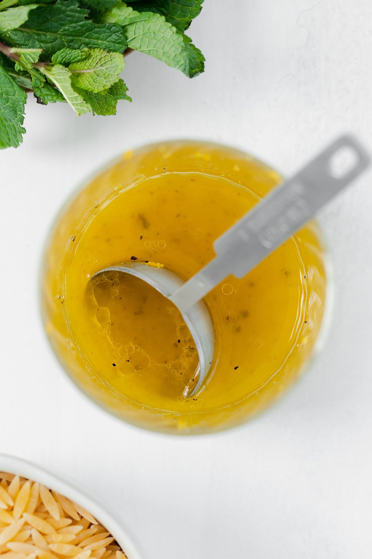 Lemon vinaigrette in a small glass with a measuring spoon in it. On a white background with fresh mint in the top left corner and dry orzo in the bottom left corner.