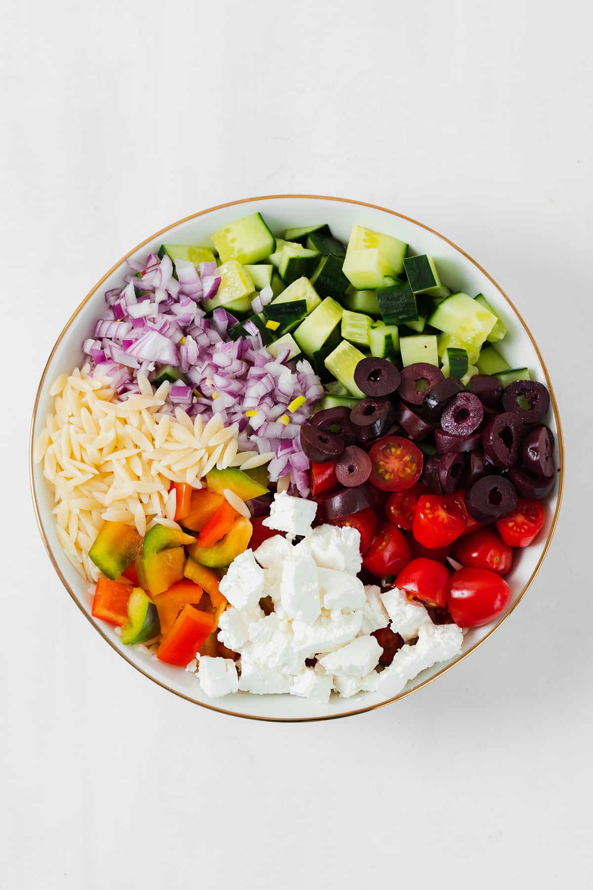 All the ingredients for the orzo salad chopped and laid out in a white bowl with a gold rim.