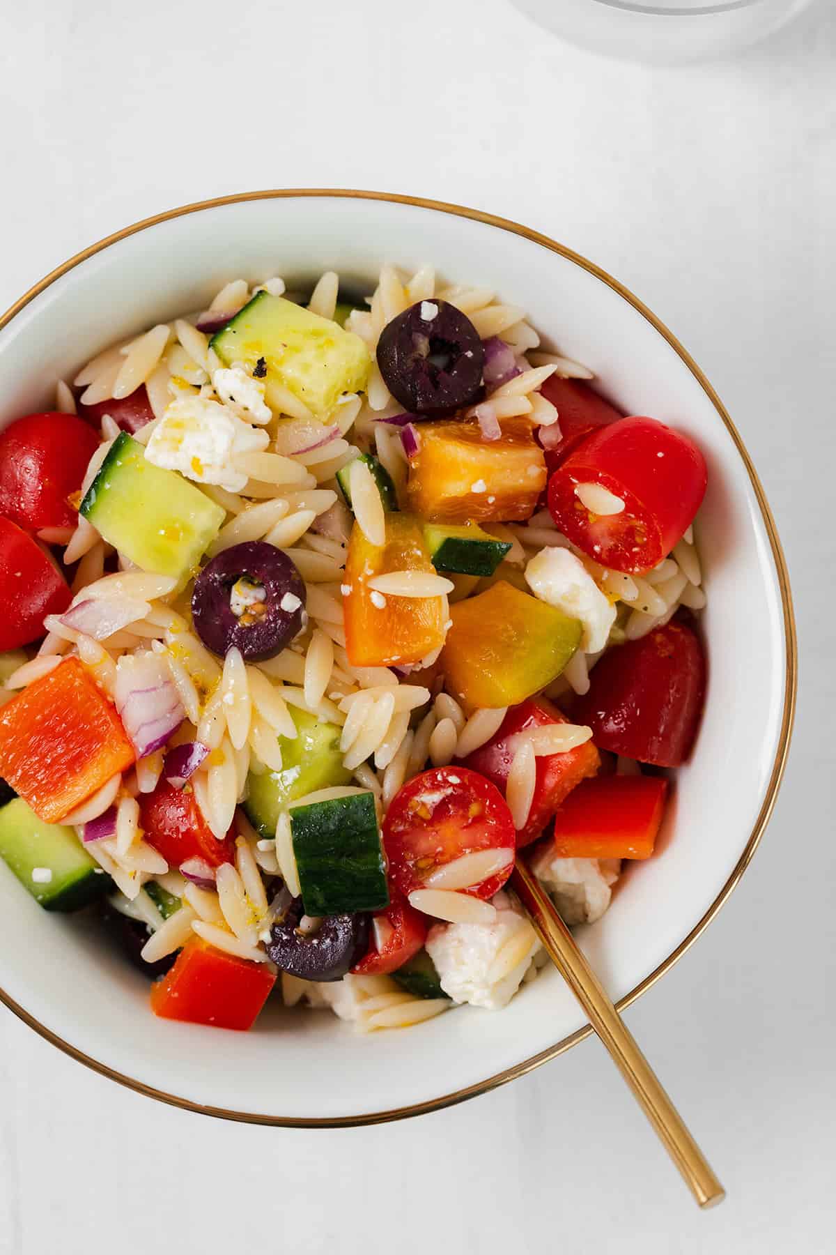 A close up of the orzo salad in a white bowl with a golden rim. On a white background.