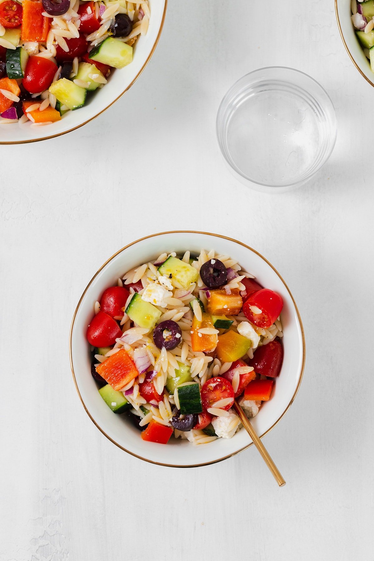 Orzo salad shown in a small white bowl with a gold rim. A small glass of water in the right side above it. Partially shown bigger bowl on the top left corner and a smaller bowl in the top right corner. Both bowls have the pasta salad in them.