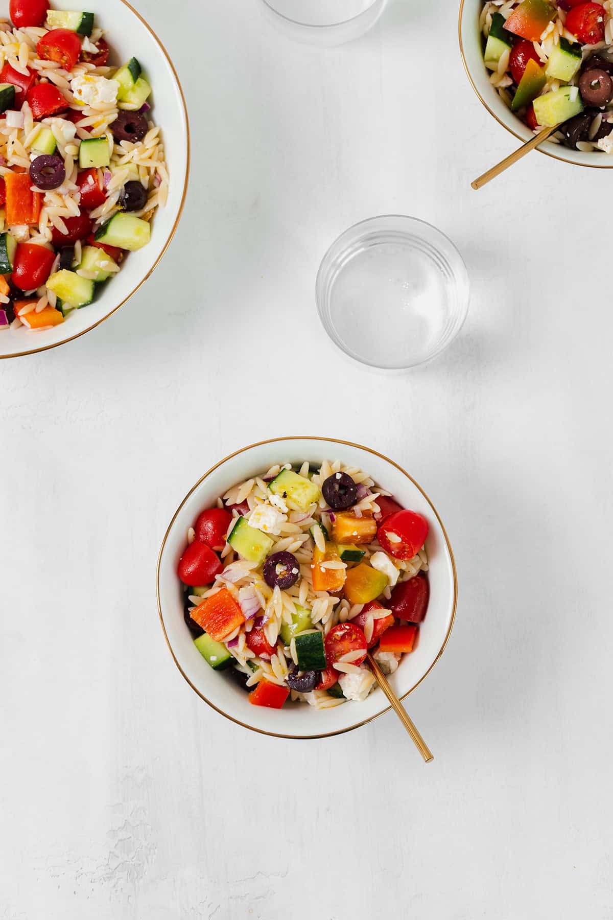 Orzo salad shown in a small white bowl with a gold rim. A small glass of water in the right side above it. Partially shown bigger bowl on the top left corner and a smaller bowl in the top right corner. Both bowls have the pasta salad in them.