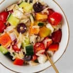 A close up of the orzo salad in a white bowl with a golden rim. On a white background.