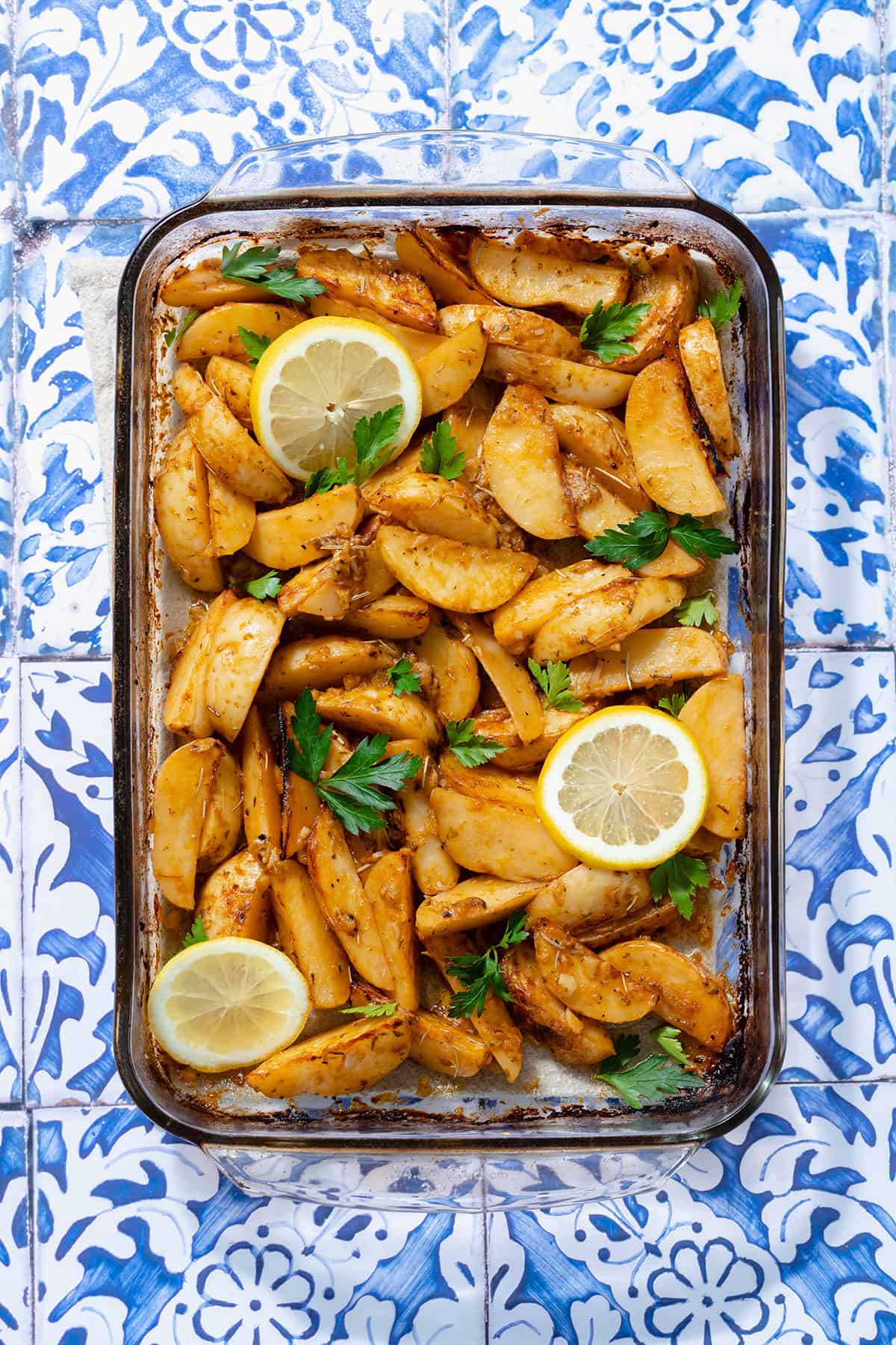 A photo of Mediterranean Potatoes garnished with three slice of lemon and chopped parsley. Blue tile background.