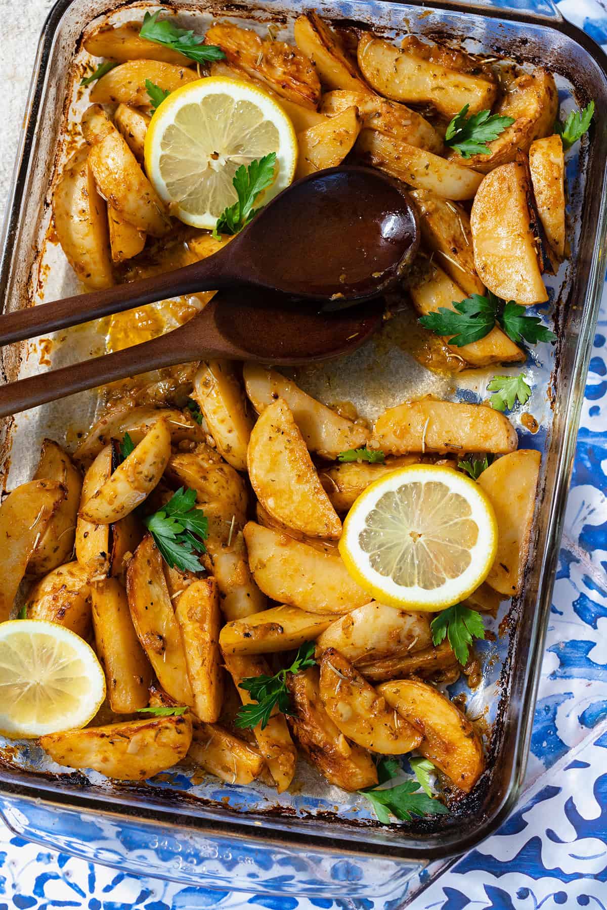 A close up photo of Mediterranean Potatoes garnished with three slice of lemon and chopped parsley. Blue tile background and two wooden spoons on the left side of the glass baking dish.