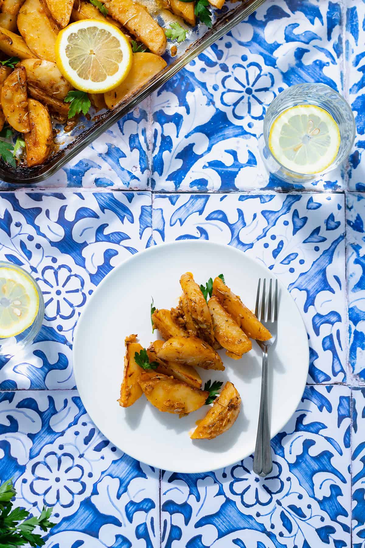 A photo of Mediterranean Potatoes garnished with three slice of lemon and chopped parsley on a white plate and blue tile background. Glass baking dish with more potatoes in the top left cover. A small glass with lemon water on the right side of the plate.