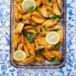 A square photo of Mediterranean Potatoes in a glass baking dish on blue tile background.