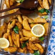 A close up photo of Mediterranean Potatoes garnished with three slice of lemon and chopped parsley. Blue tile background and two wooden spoons on the left side of the glass baking dish.