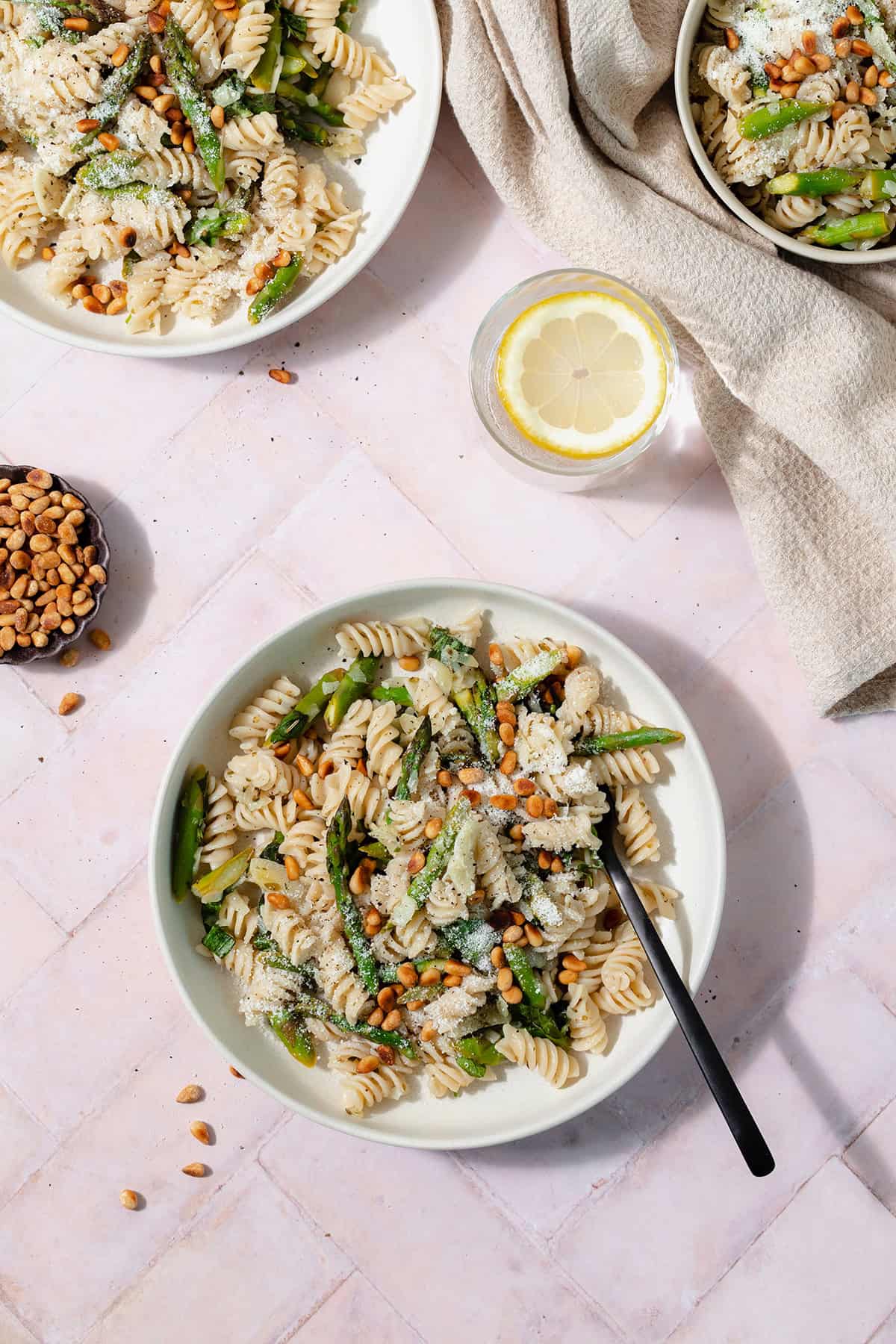 A low light green bowl with asparagus pasta and a black fork on a light pink tile background. Two more bowls, a little bowl with toasted pine nuts, and a glass of water with a lemon slice in the background.