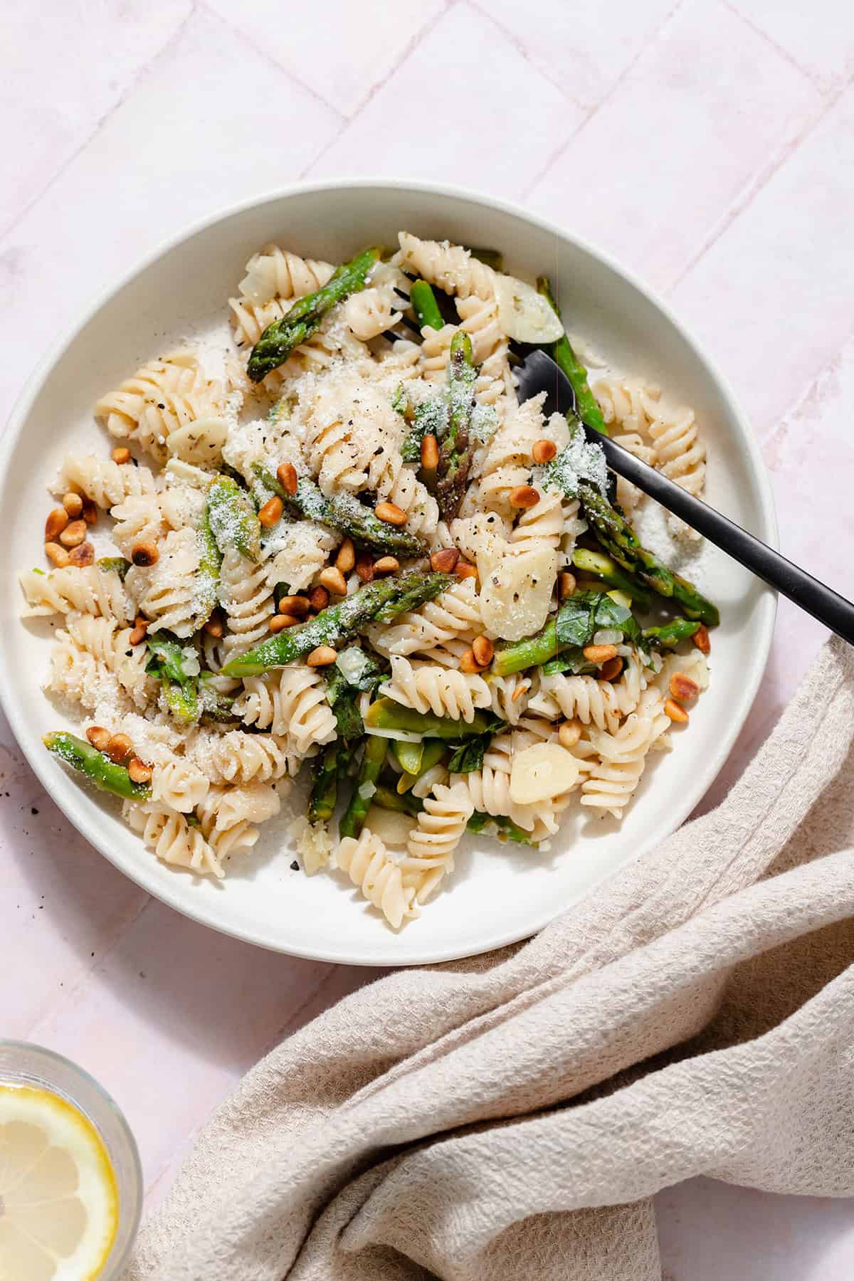 Asparagus pasta with toasted pine nuts and fresh basil on a light green low bowl with a black fork in the bowl. On a light pink tile background.