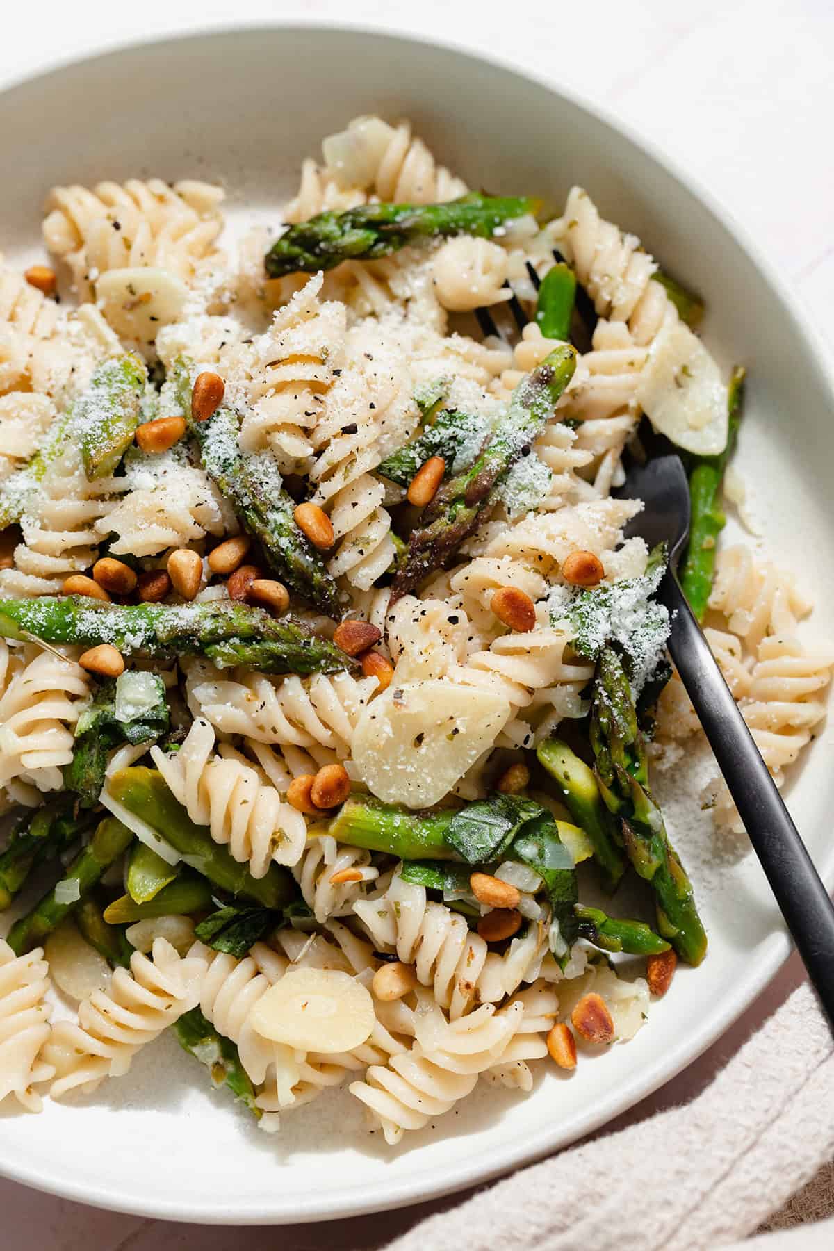 A close up of Asparagus pasta with toasted pine nuts and fresh basil on a light green low bowl with a black fork in the bowl. On a light pink tile background.