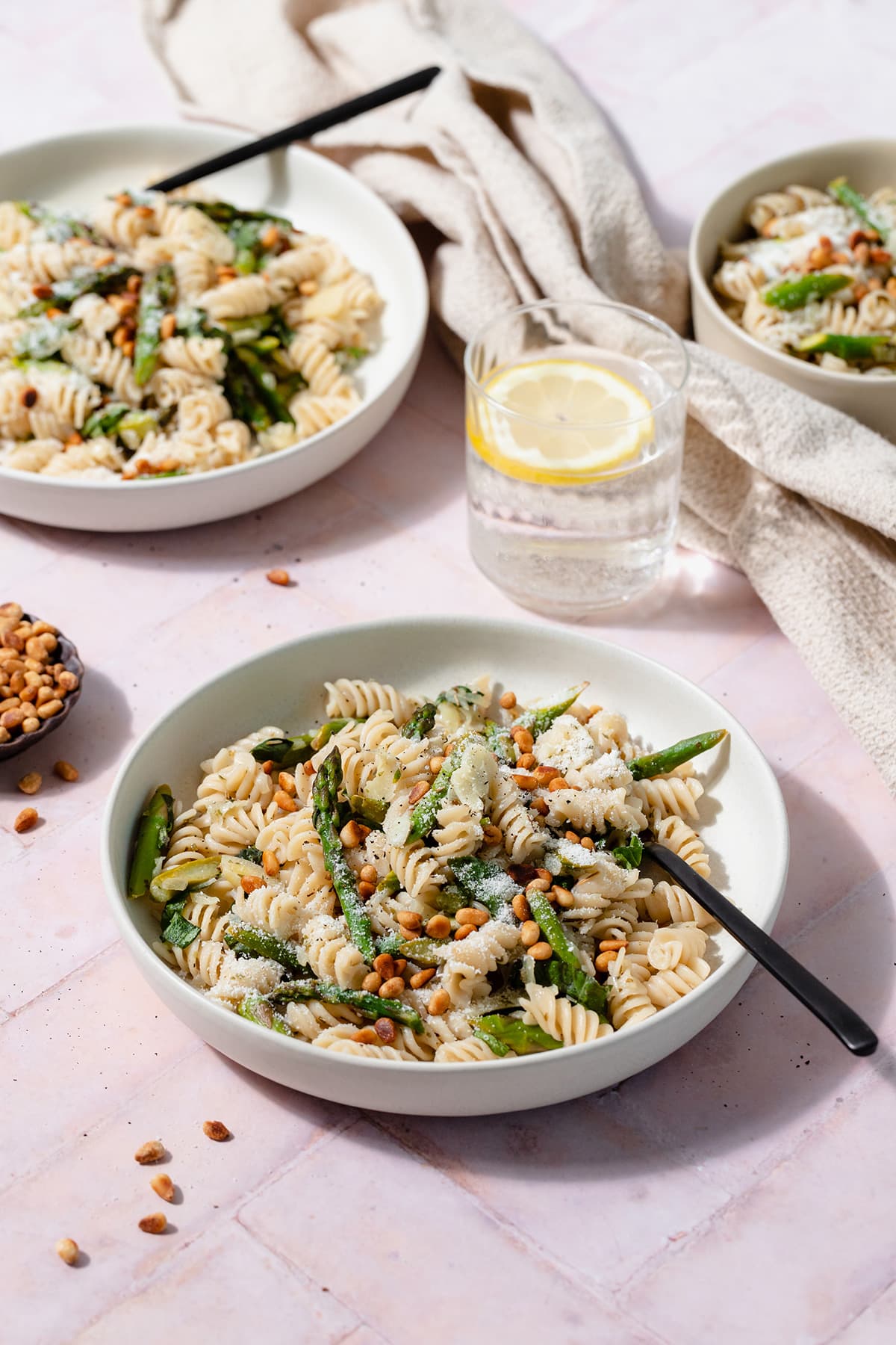 A low light green bowl with asparagus pasta and a black fork on a light pink tile background. Two more bowls, a little bowl with toasted pine nuts, and a glass of water with a lemon slice in the background.