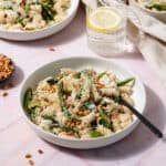 A low light green bowl with asparagus pasta and a black fork on a light pink tile background. Two more bowls, a little bowl with toasted pine nuts, and a glass of water with a lemon slice in the background.