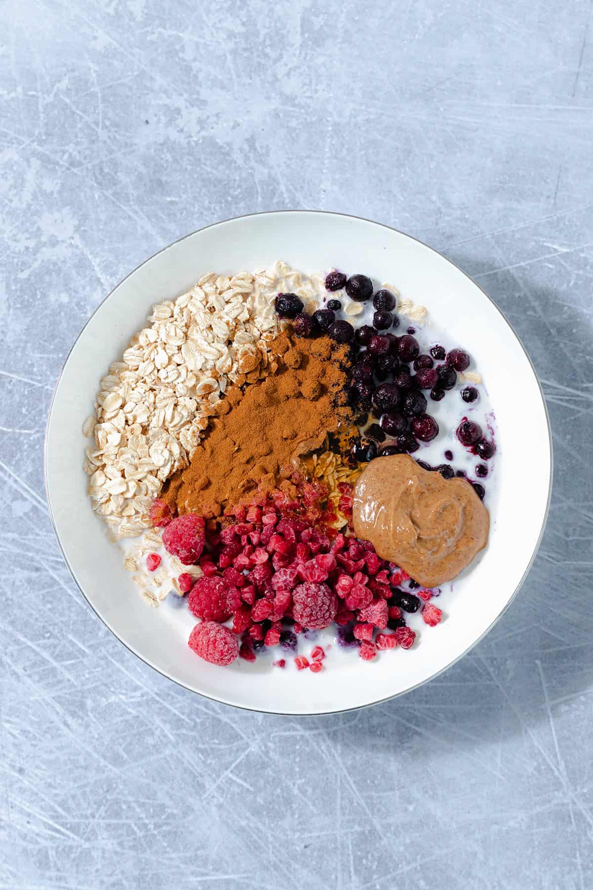 All the ingredients for blueberry oats in a white shallow bowl on a metal background.