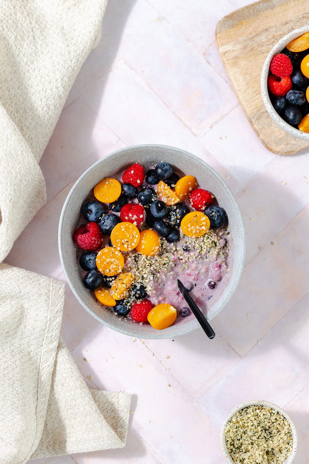 Overhead shot of blueberry overnight oats in a grey bowl topped with blueberries, raspberries, golden berries, and hemp seeds. Pink tile background.