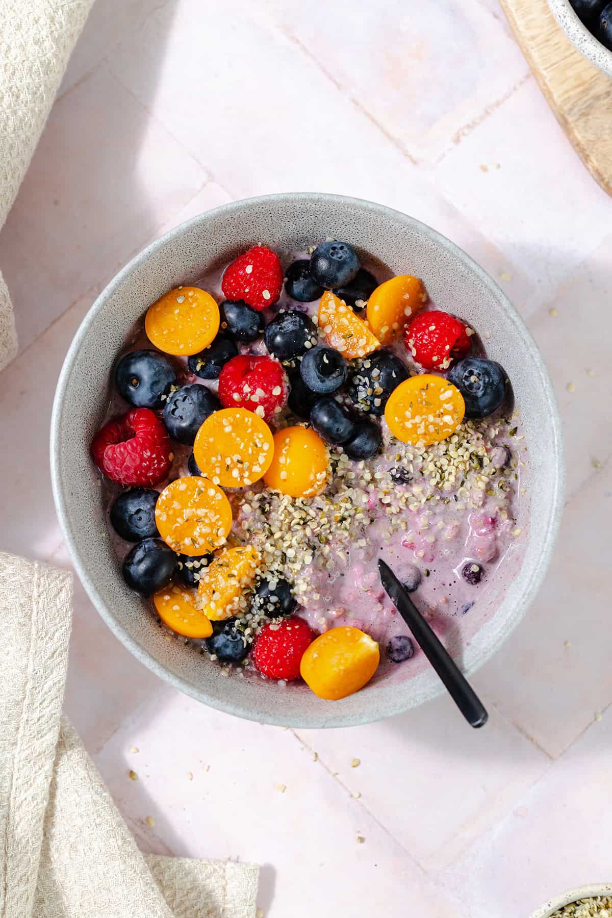 Overhead shot of blueberry overnight oats in a grey bowl topped with blueberries, raspberries, golden berries, and hemp seeds. Pink tile background.