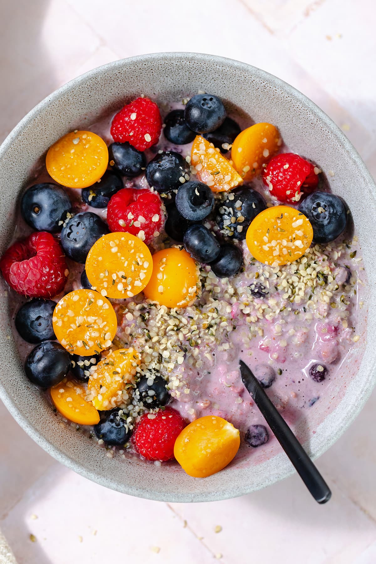 Overhead shot of blueberry overnight oats in a grey bowl topped with blueberries, raspberries, golden berries, and hemp seeds. Pink tile background.