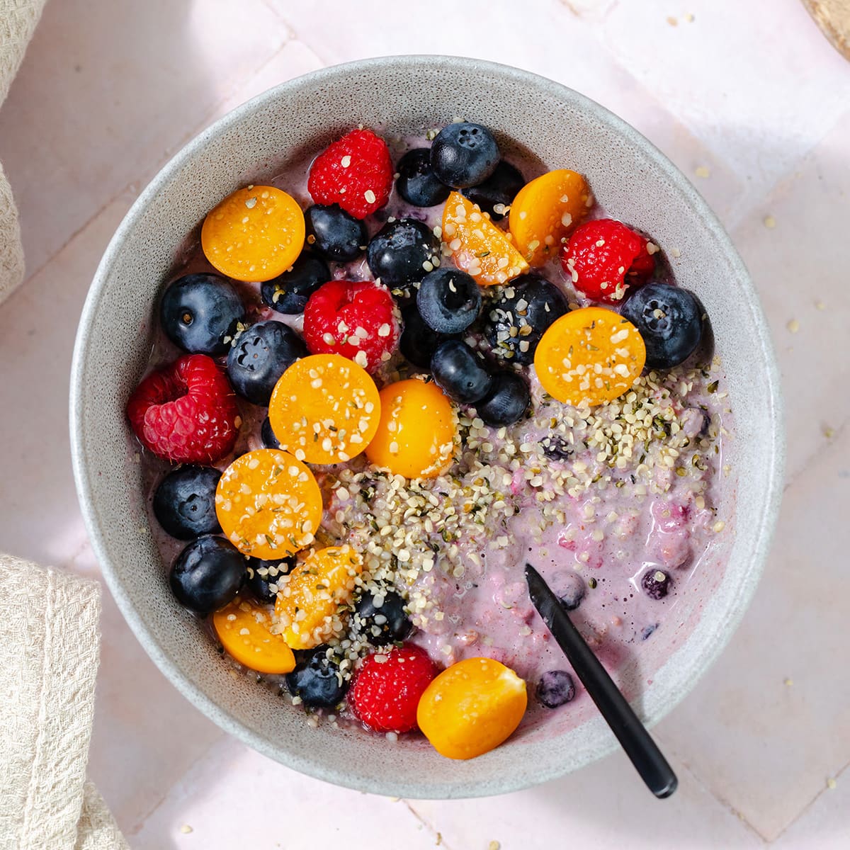 Overhead shot of blueberry overnight oats in a grey bowl topped with blueberries, raspberries, golden berries, and hemp seeds. Pink tile background.