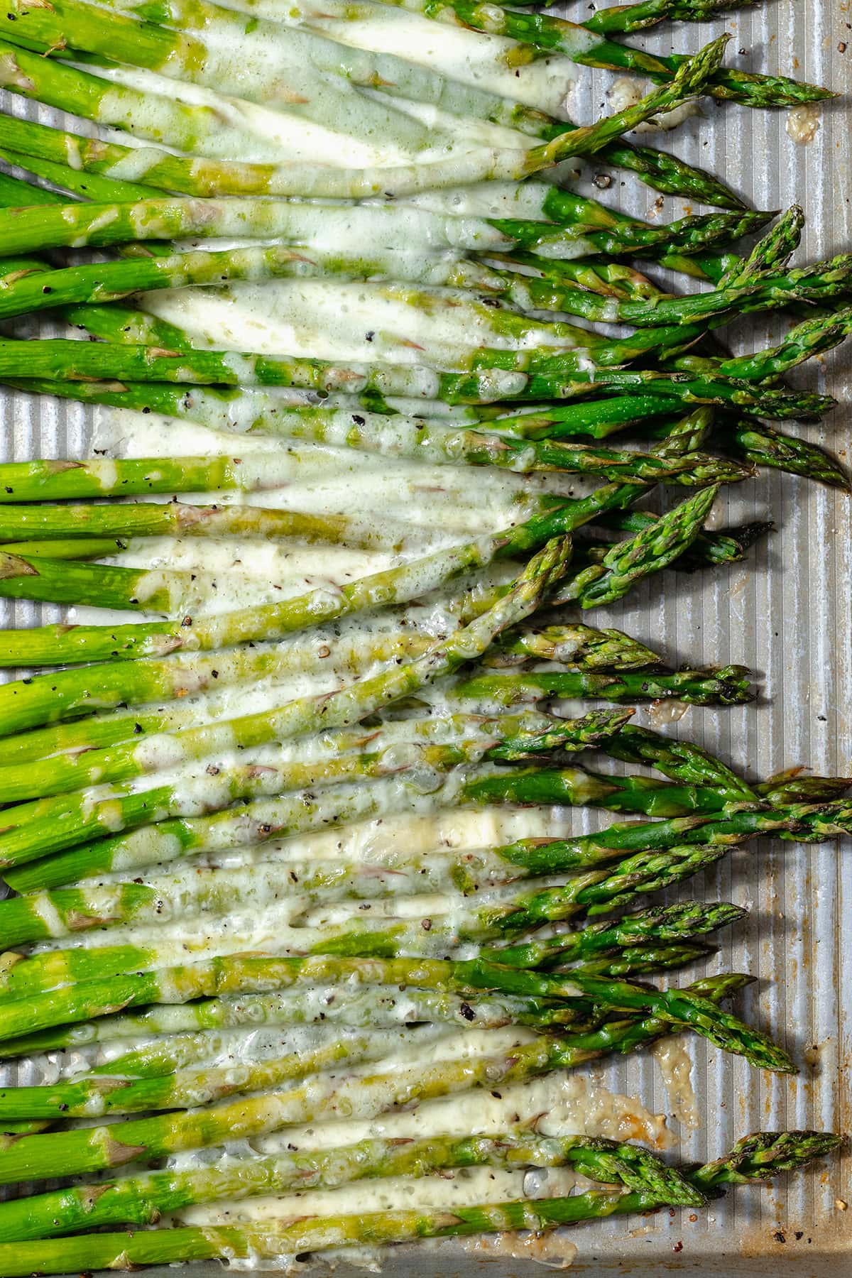A close up photo of roasted asparagus spears on a baking sheet with cheese sprinkled on that has melted in the oven.