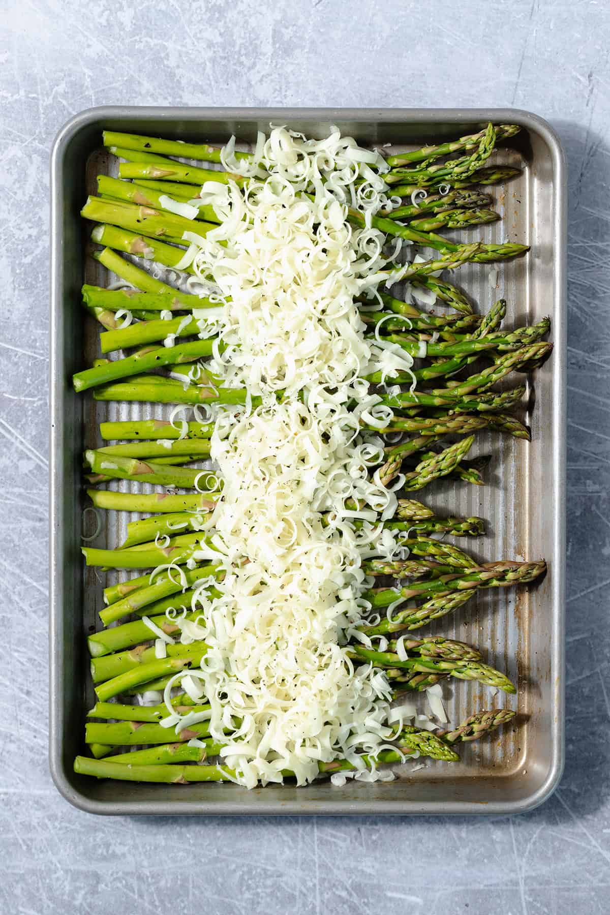 Fresh asparagus on a sheet pan sprinkled with grated cheese. Pan is on a scratched metal surface.