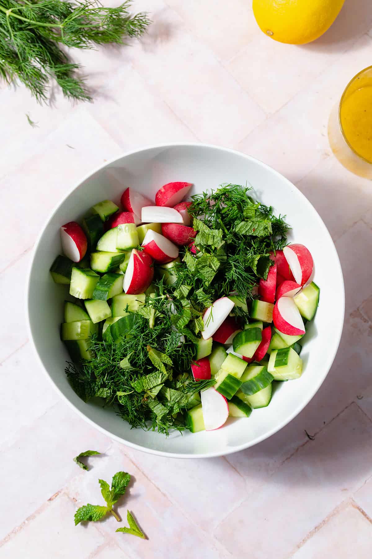 All ingredients for the cucumber radish salad in a white bowl. Chopped radishes, cucumnber, and fresh herbs. Lemon vinaigrette in a glass in the top right corner of the frame. Light pink tile table.